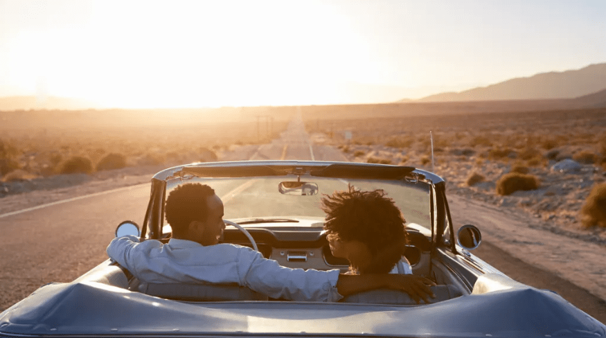 Rear view of couple on road trip driving classic convertible car towards sunset.